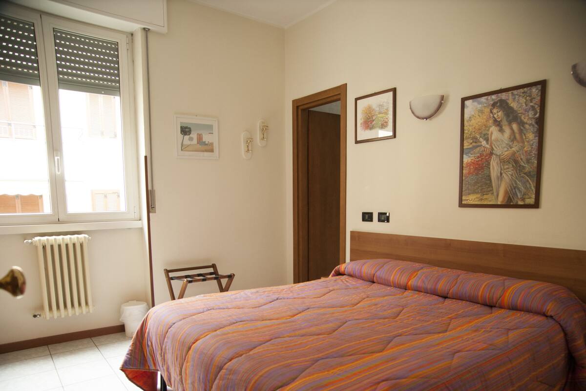 A bedroom with a bed, a radiator, a window with blinds, a door, and wall-mounted pictures.
