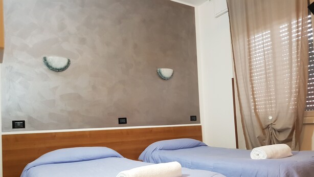 Une chambre d’hôtel avec deux lits, chacun recouvert d’une couverture bleue et agrémenté d’une serviette blanche. La chambre est dotée d’une tête de lit en bois, d’un mur à texture et d’une fenêtre avec des rideaux.