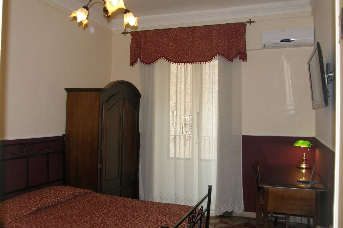A bedroom with a bed, a wooden wardrobe, a desk with a lamp, and a window with curtains.