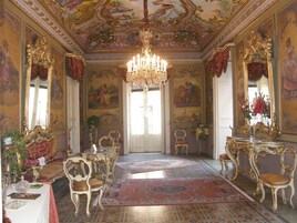 Um quarto luxuoso com móveis elaborados, um candelabro e murais detalhados nas paredes.