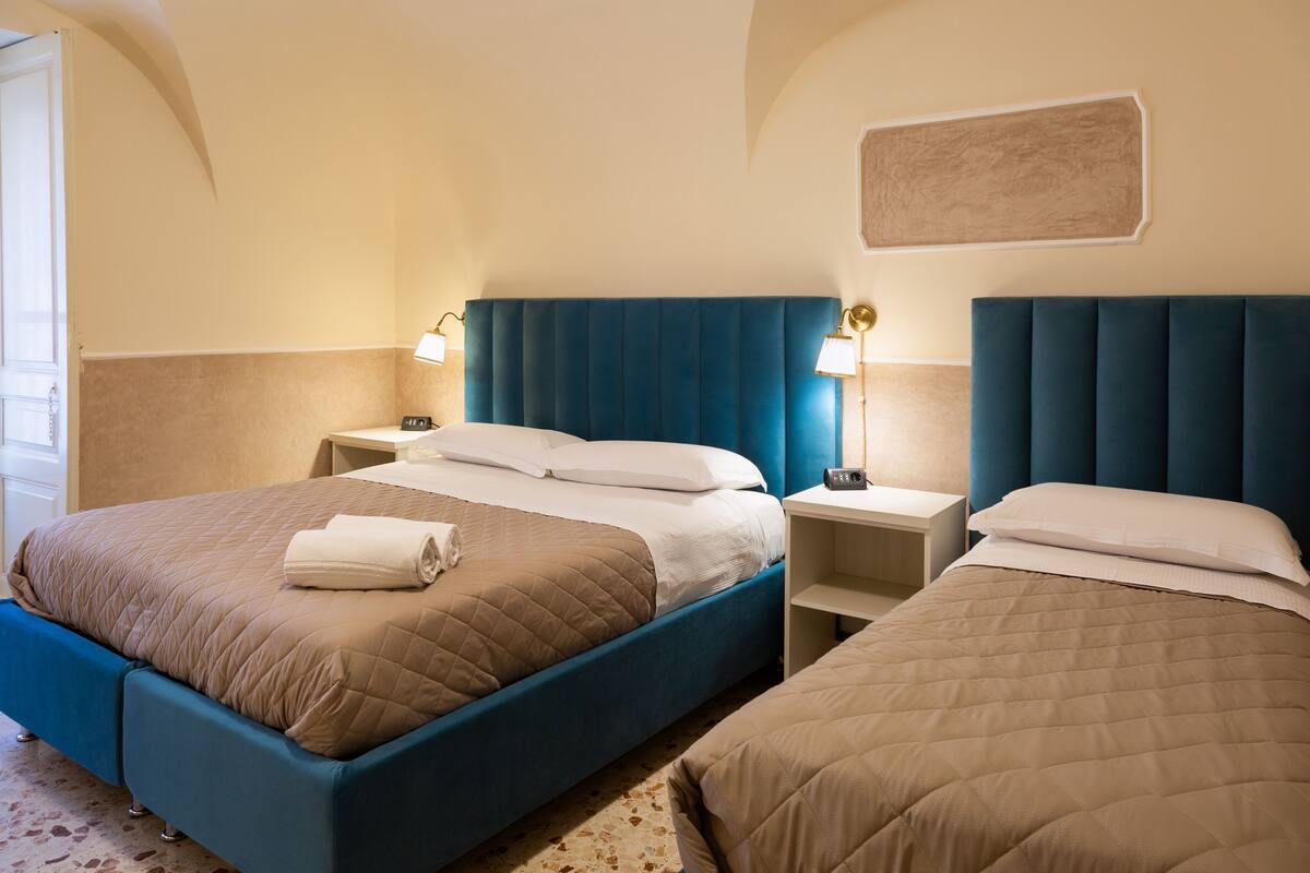 A hotel room with two beds, a blue headboard, bedside lamps, and a small nightstand.