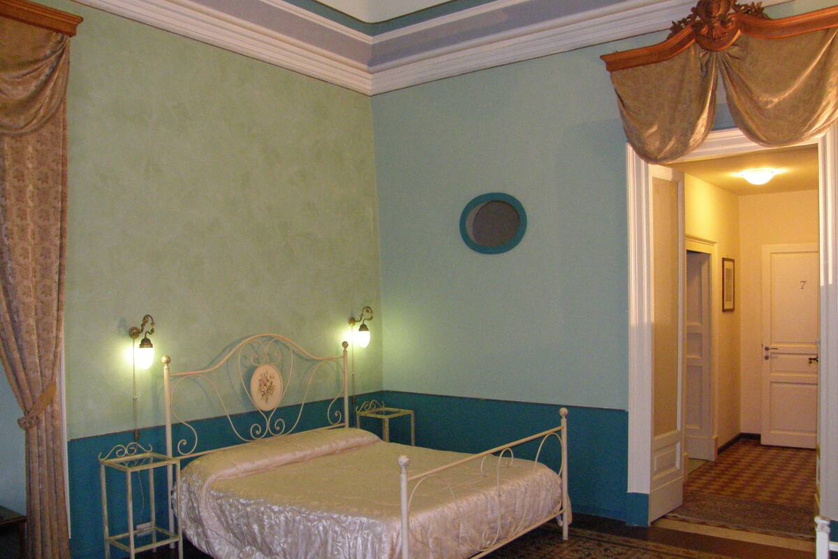 A bedroom with a single bed, a nightstand, a wall-mounted lamp, and a door leading to another room.