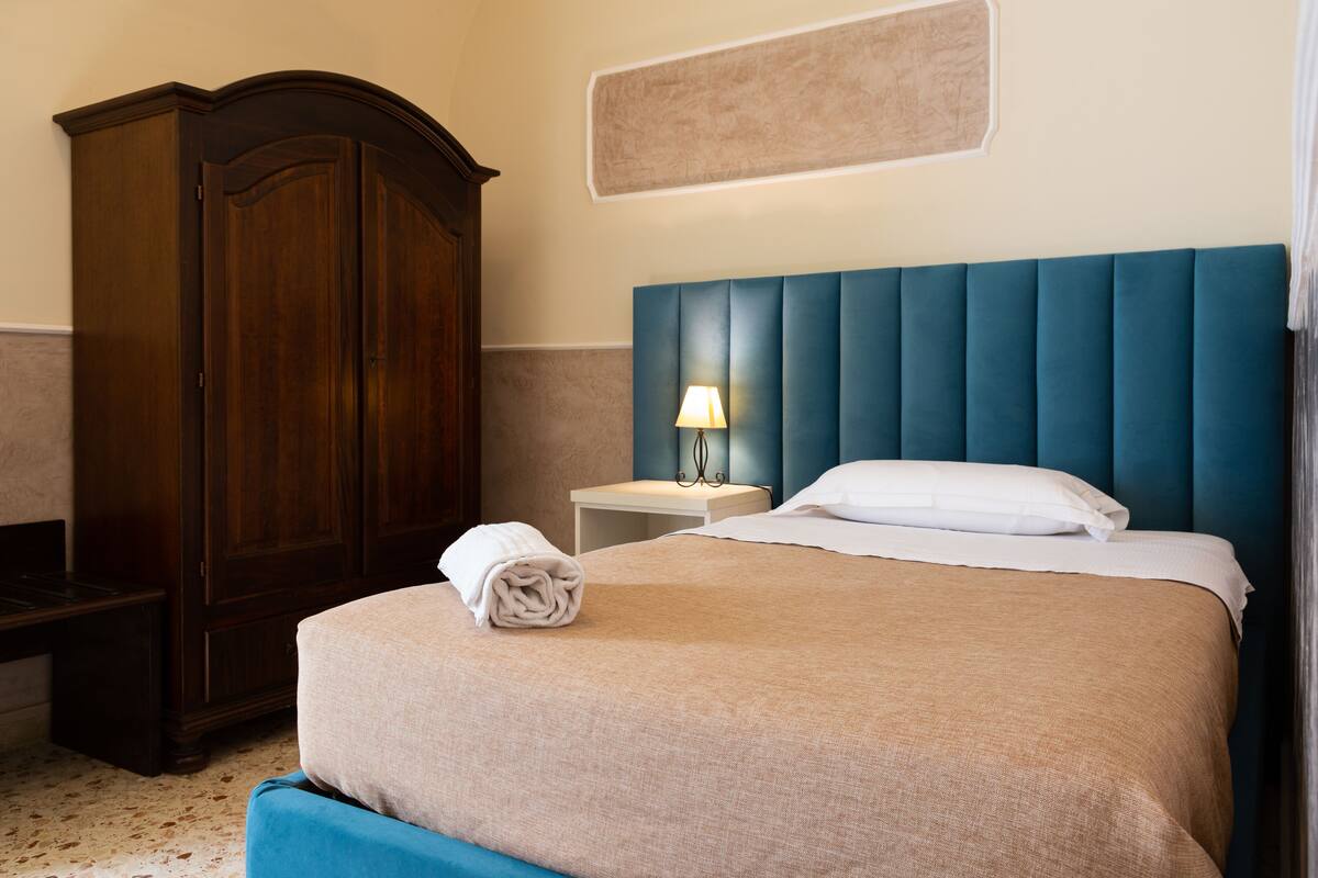 A neatly made bed with a blue headboard, a wooden wardrobe, and a small bedside table with a lamp.