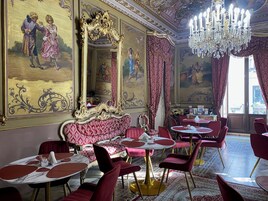 Uma área de refeições luxuosa, com espelhos dourados ornamentados, sofás de veludo vermelho e cadeiras vermelhas. As mesas são redondas com tampos de mármore branco.