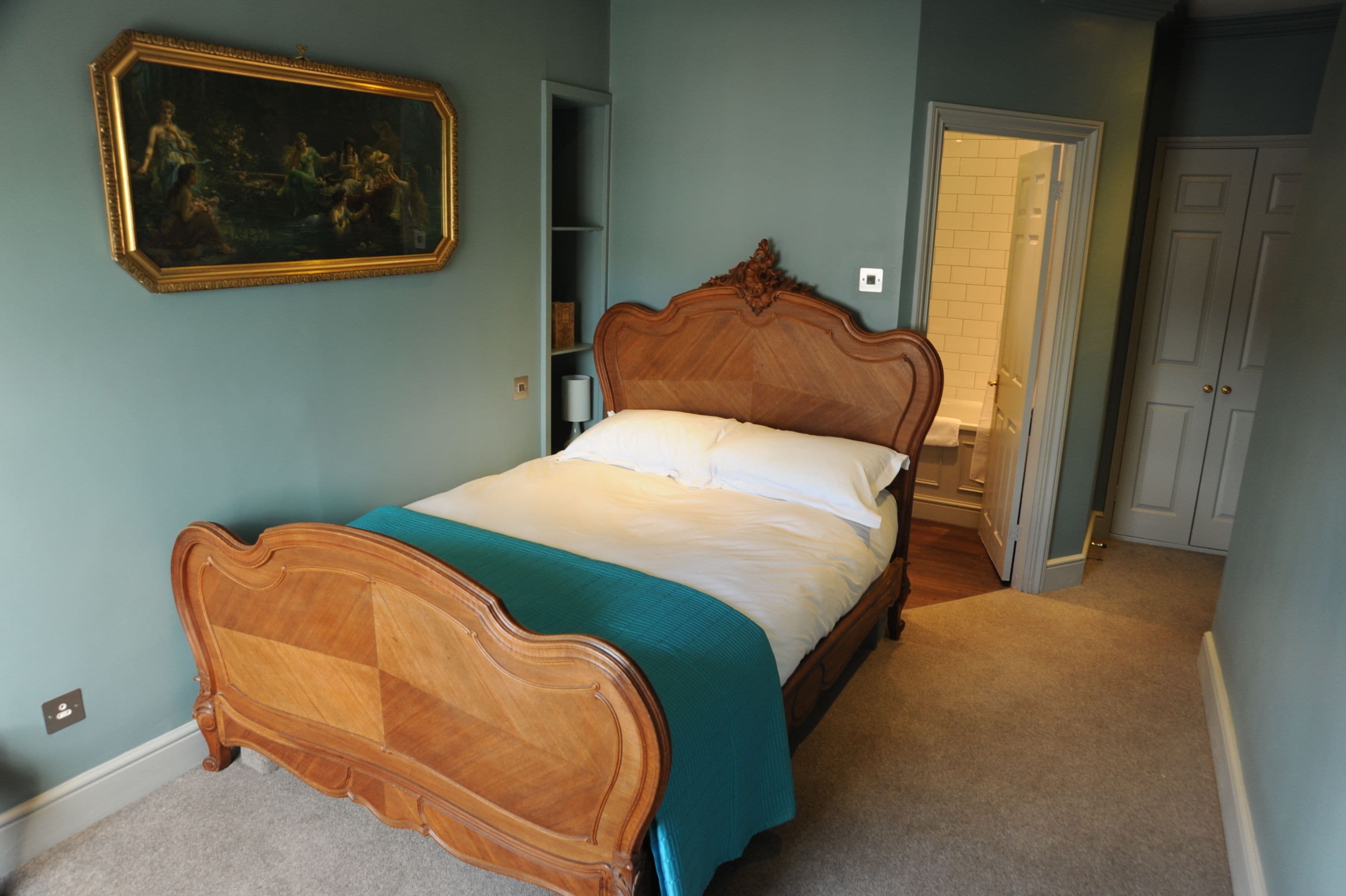 A bedroom with a wooden bed, a teal bedspread, and a classic painting on the wall.