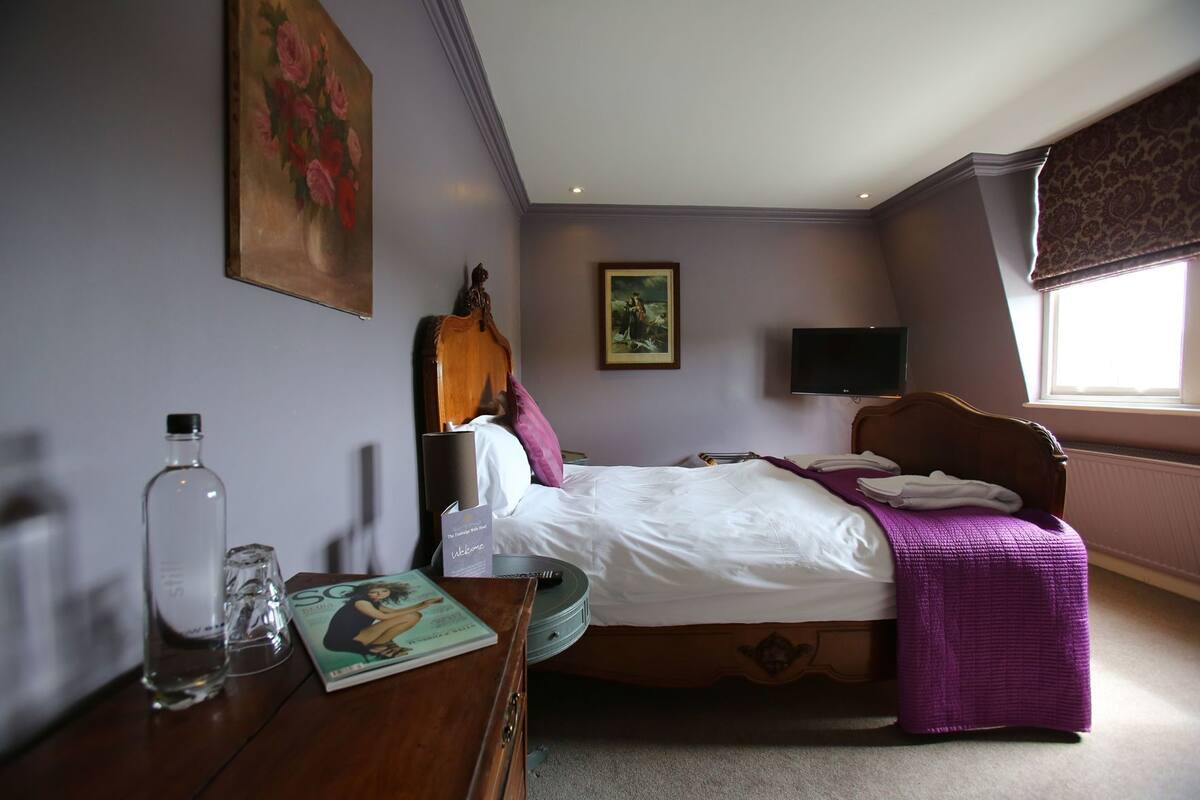 A hotel room with a bed, a bedside table with a magazine, a bottle, and a painting on the wall.