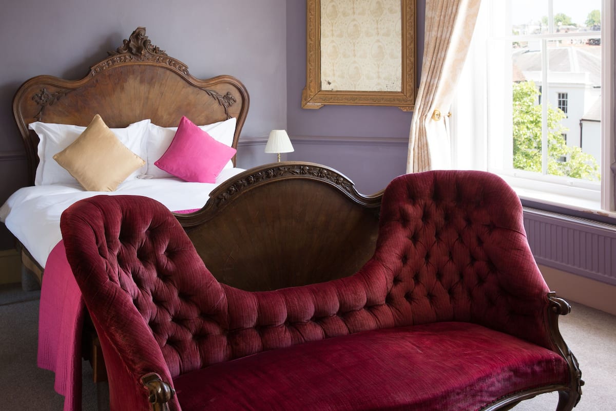 A room with a bed, a red velvet chair, a nightstand with a lamp, and a window with a view of buildings.
