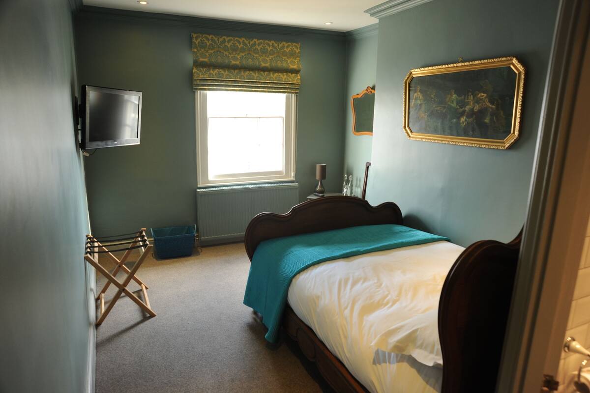 A bedroom with a bed, a television, a window with a curtain, a painting on the wall, and a radiator.