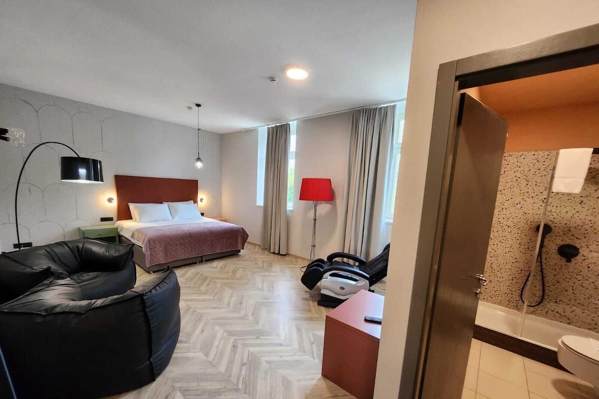 A hotel room with a bed, a desk, a chair, a massage chair, a lamp, and a bathroom with a shower and toilet.