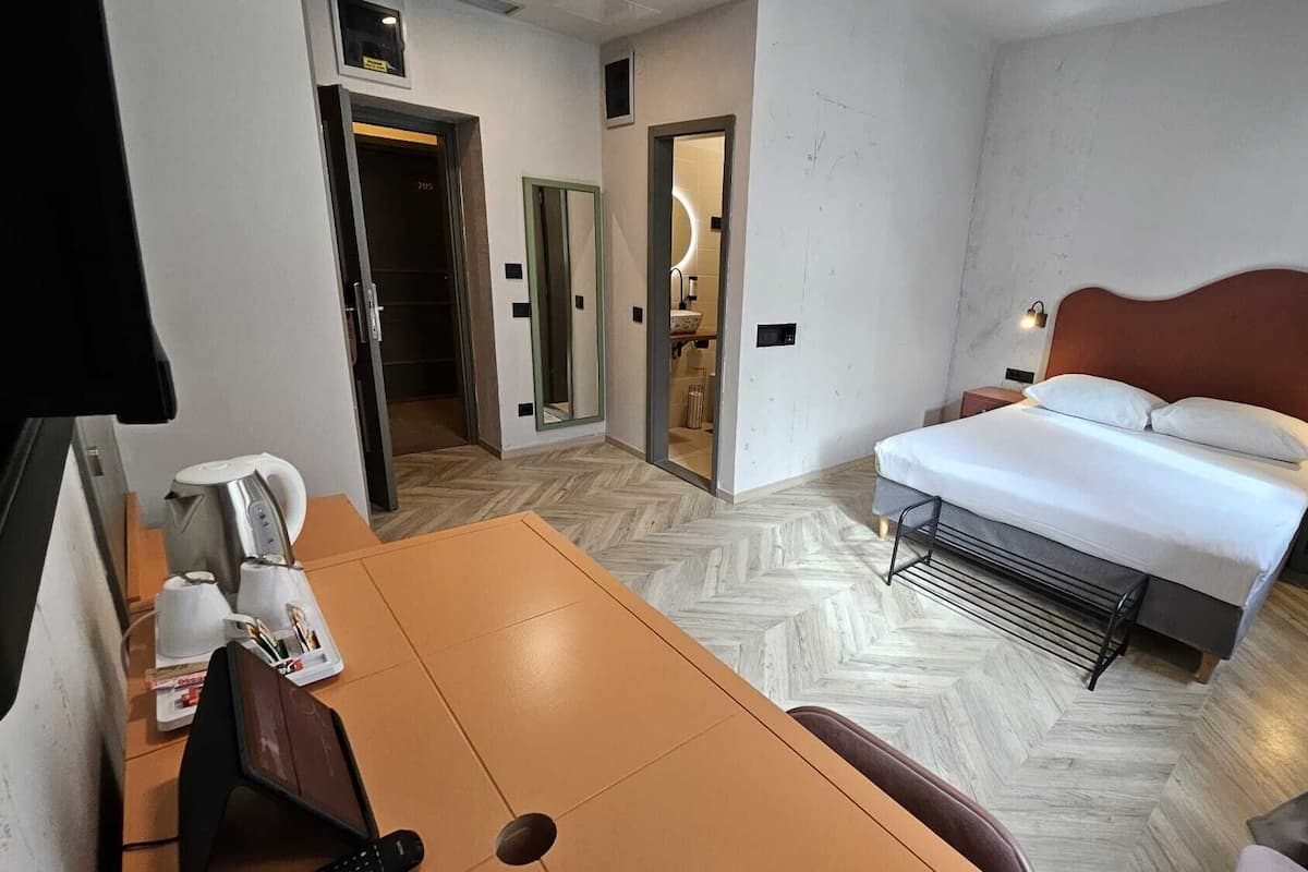 A hotel room with a bed, a desk with a kettle, a chair, and a bathroom visible through an open door.