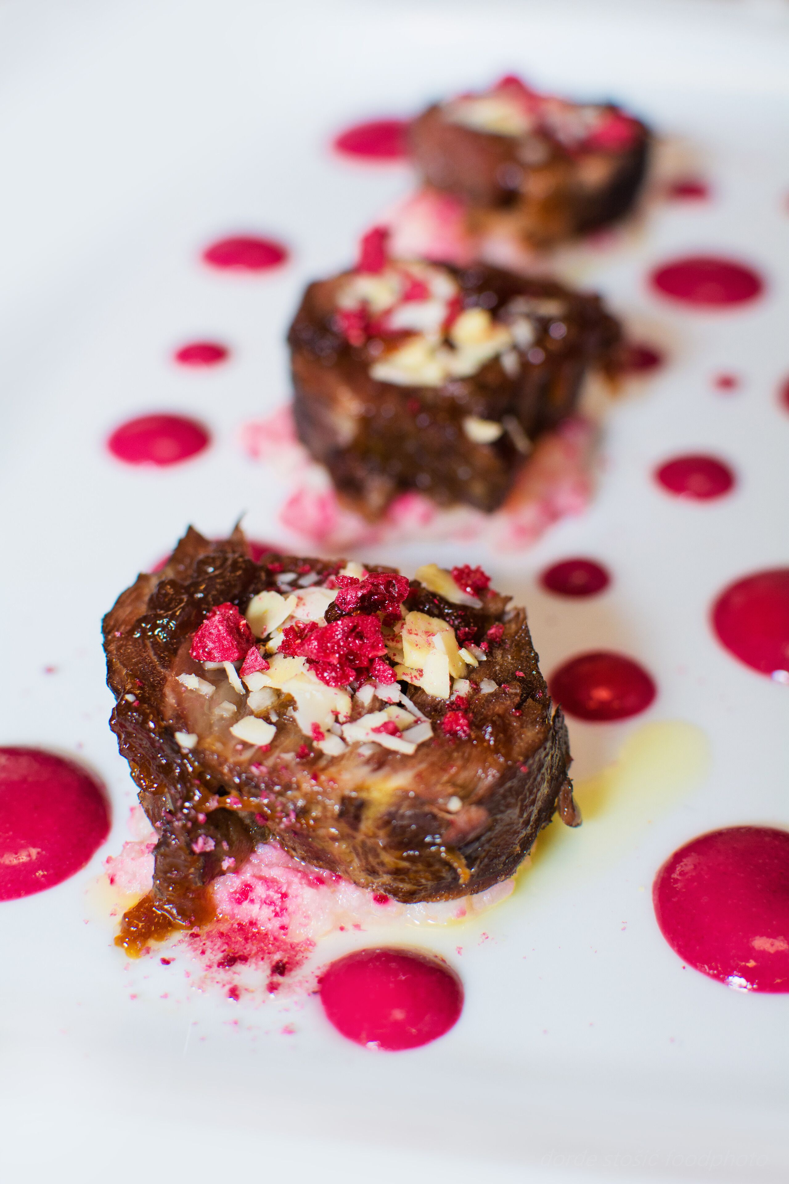A dessert with a chocolate base, topped with fruit and nuts, served on a white plate with pink sauce.