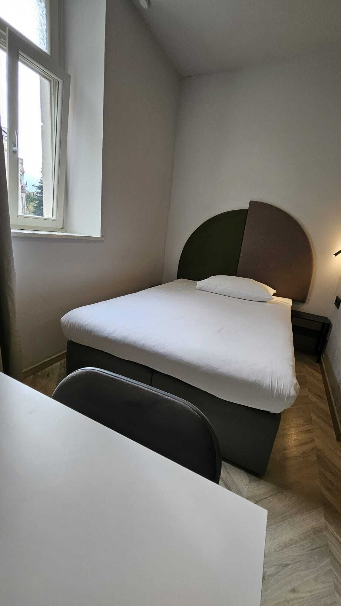 A small, neatly arranged bedroom with a single bed, a bedside table, a chair, and a window.