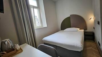 Una habitación de hotel pequeña y ordenada, con una cama, una mesita de noche, una silla, una ventana con cortinas y vistas a edificios.
