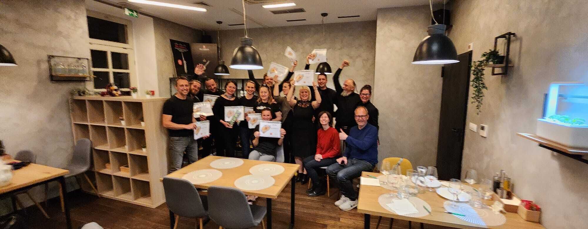 Un grupo de personas en un comedor moderno con mesas dispuestas para una comida, estantes con diversos objetos y una lámpara colgante grande.