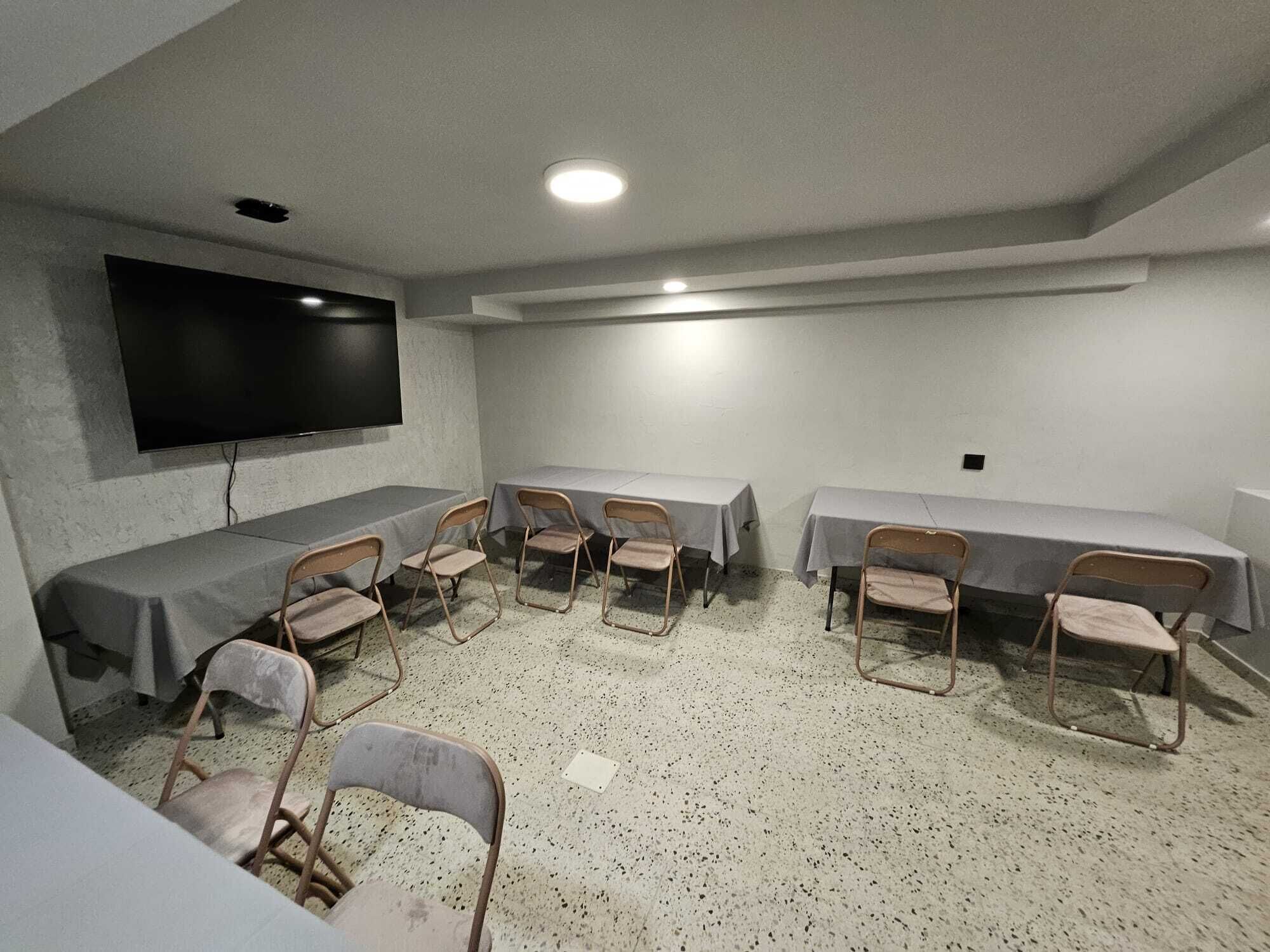Un salón de conferencias con una gran pantalla plana, varias mesas con sillas y un suelo moteado.