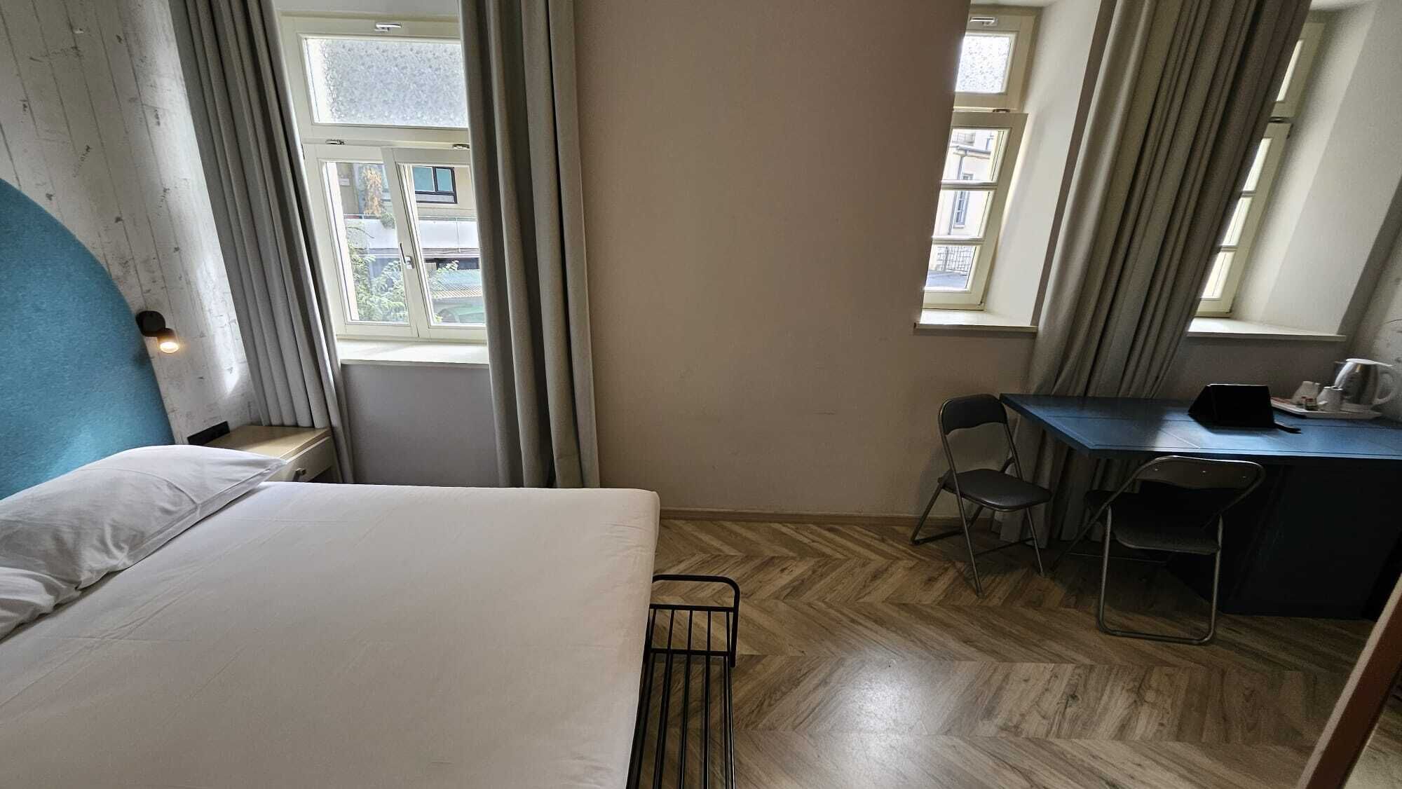 A small, compact hotel room with a single bed, a desk, two chairs, and a window with curtains.