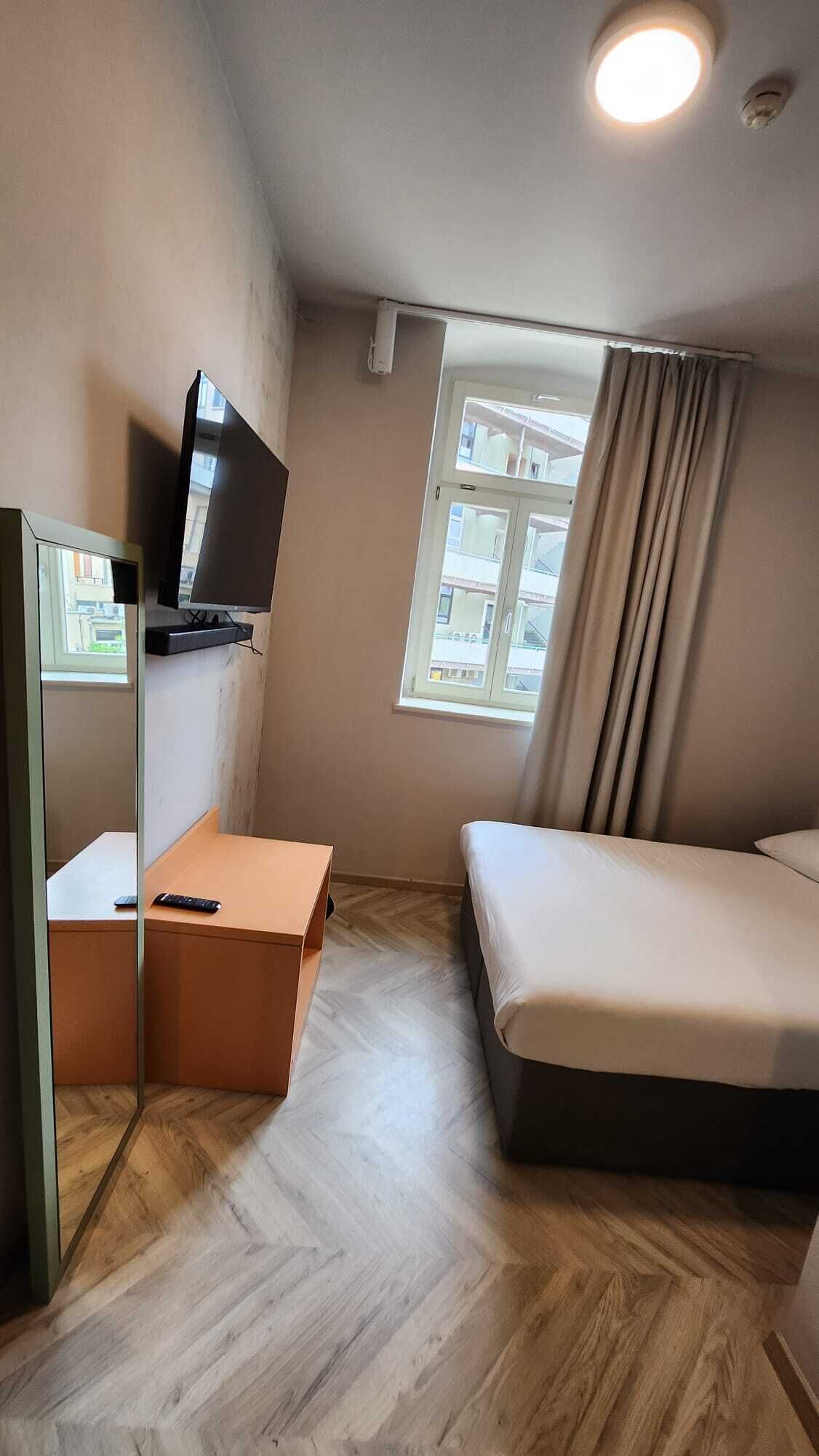 A compact hotel room with a bed, a desk with a television, a window with curtains, and a view of buildings outside.