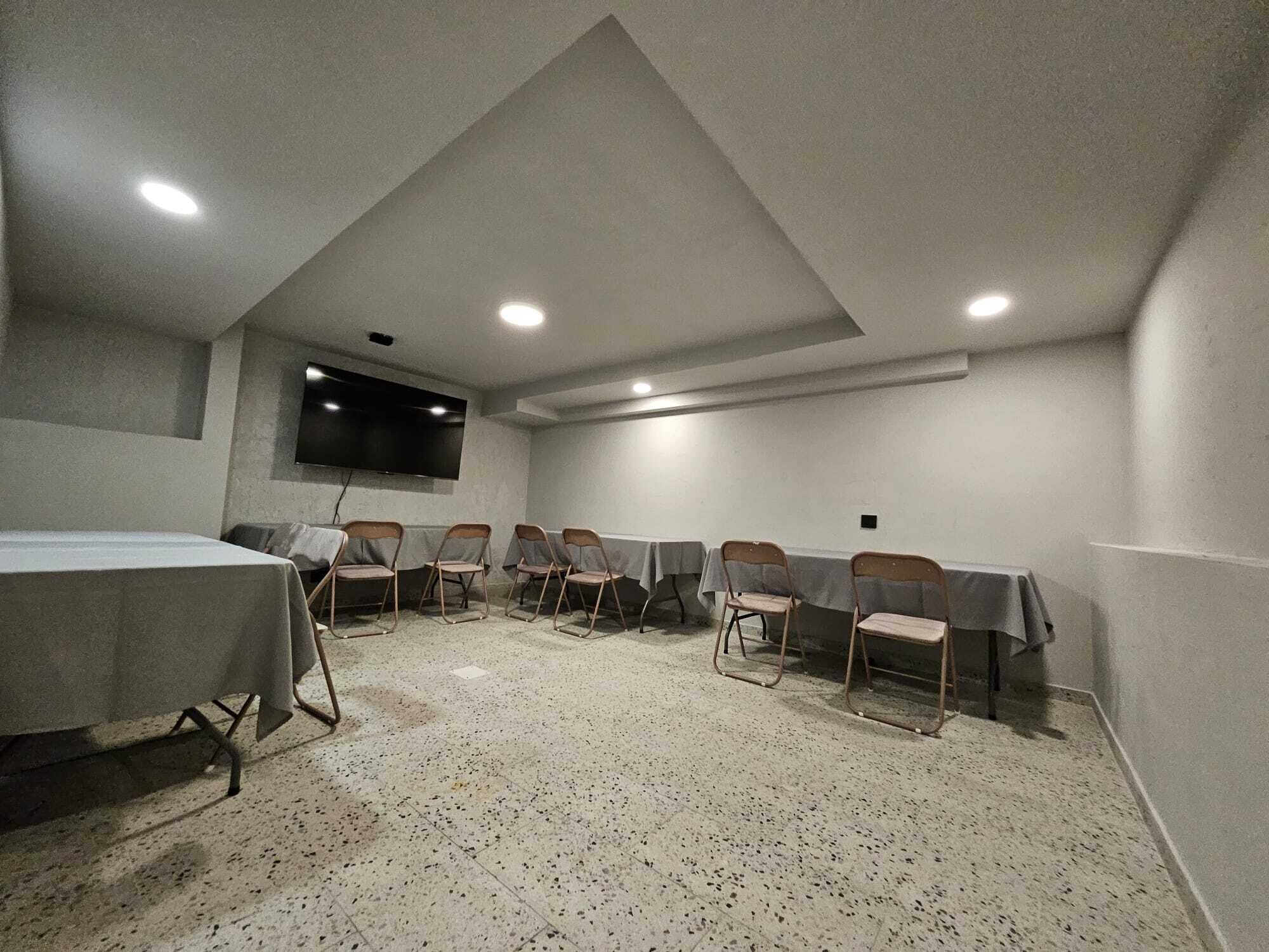 A conference room with a high ceiling, recessed lighting, a flat-screen TV, and multiple tables with chairs.