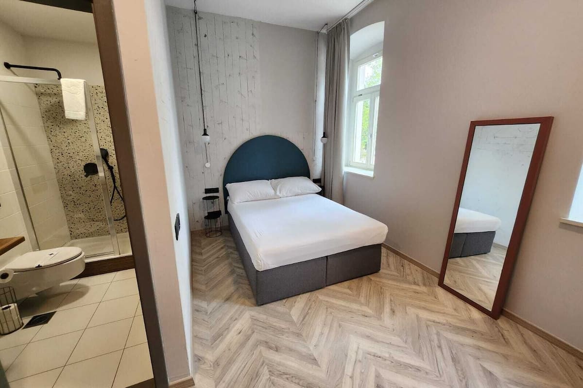 A bedroom with a bed, a mirror, a window, and a shower area.