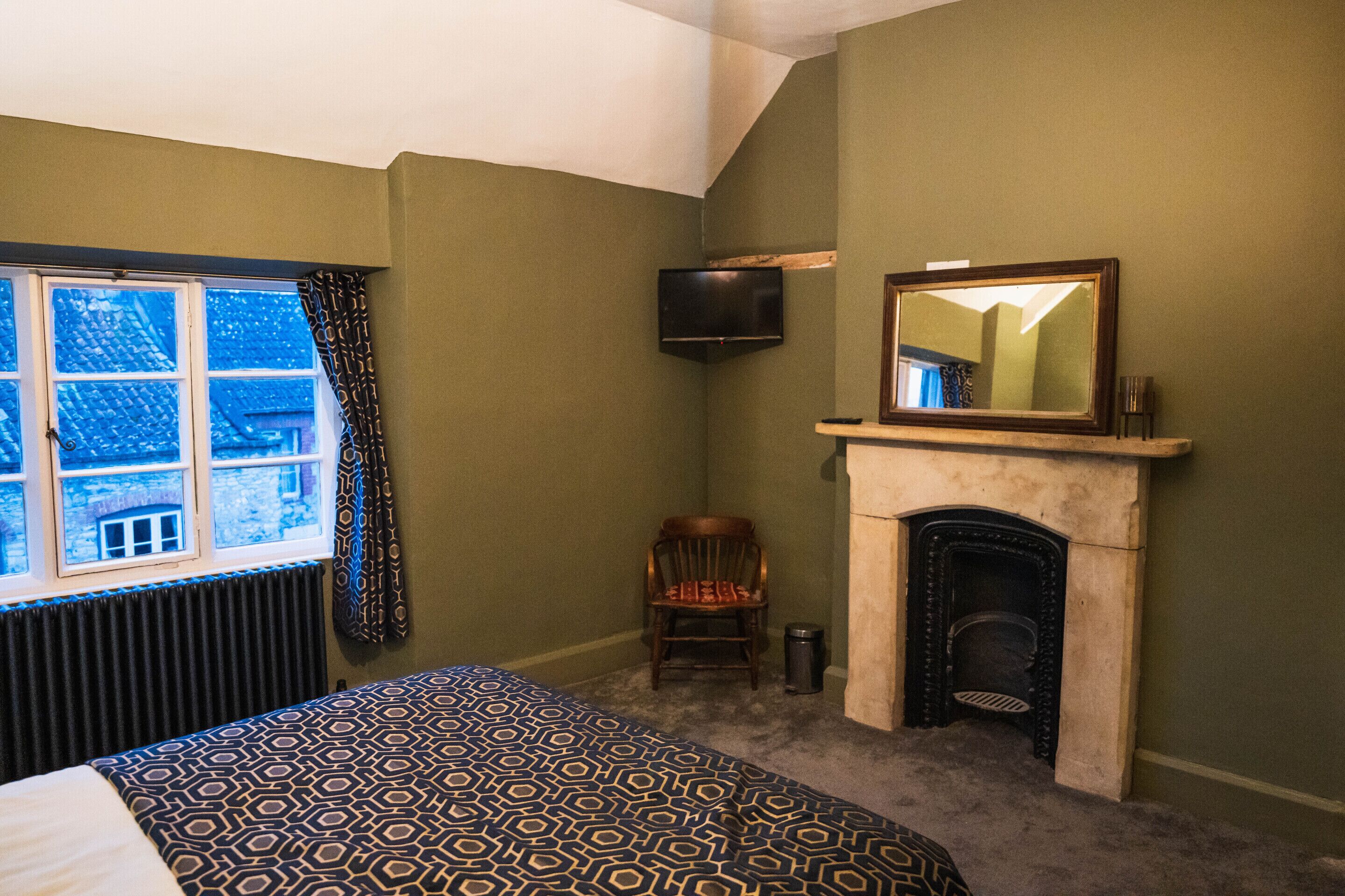 A bedroom with a patterned bedspread, a fireplace, a television mounted on the wall, and a window with curtains.