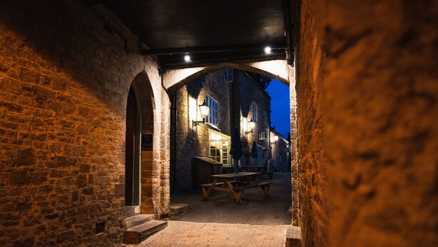 Une ruelle étroite aux murs de pierre, avec un banc en bois et une table entourée de chaises.