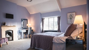 Une chambre à coucher comprenant un lit, une table de chevet, une lampe, une chaise, un bureau, une cheminée et un téléviseur fixé au mur.