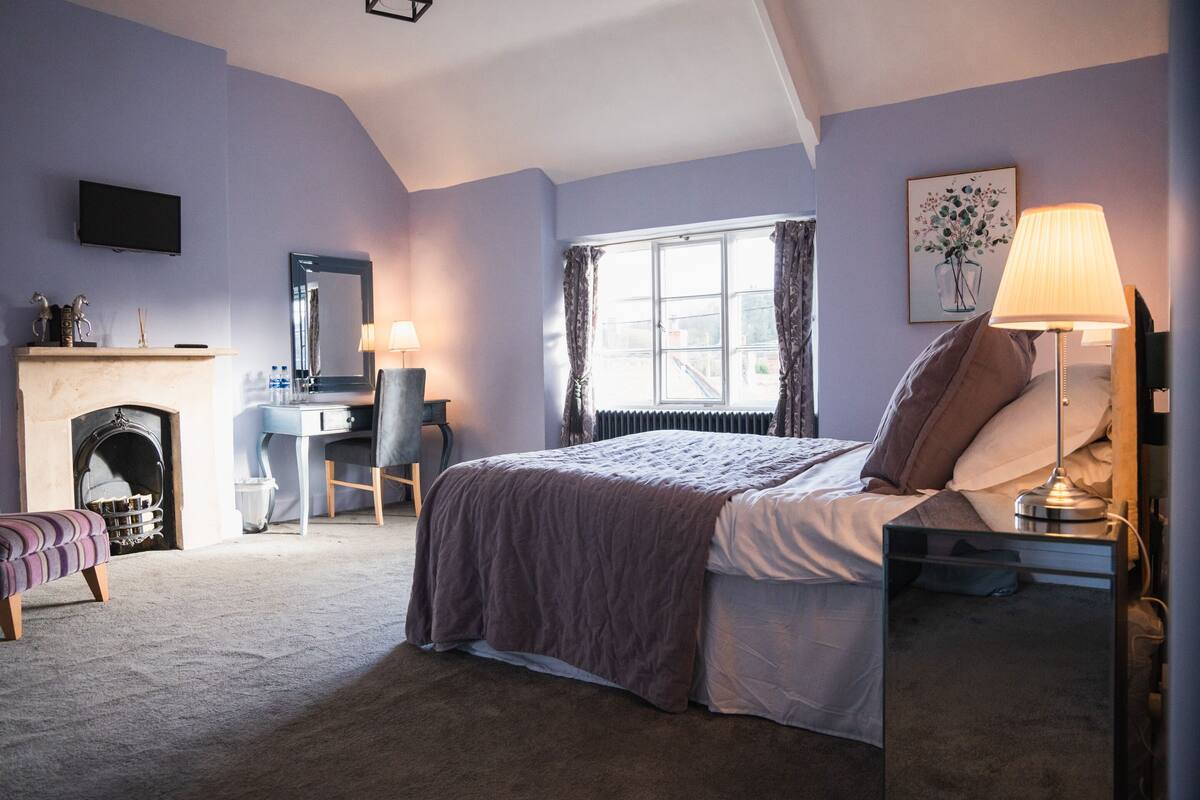 A bedroom with a bed, bedside table, lamp, chair, desk, fireplace, and a television on the wall.