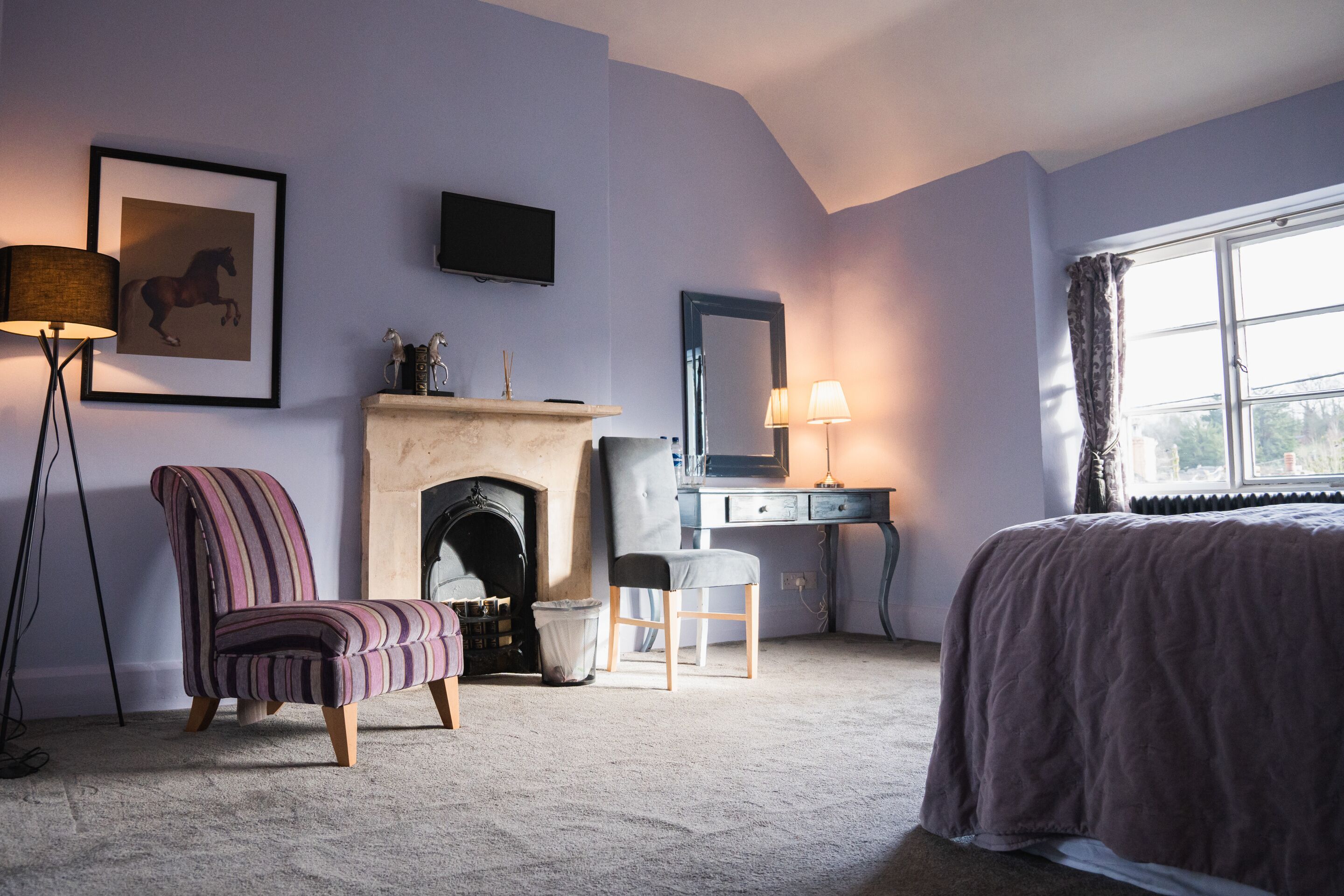 A bedroom with a bed, a chair, a desk, a fireplace, a television, and a window with curtains.