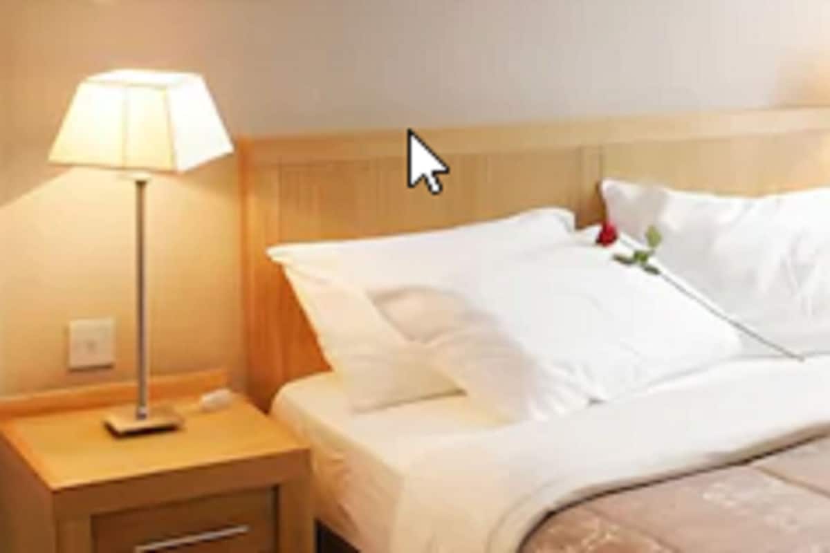 A neatly made bed with white linens and a beige comforter, two bedside lamps, a bedside telephone, and a window with curtains.