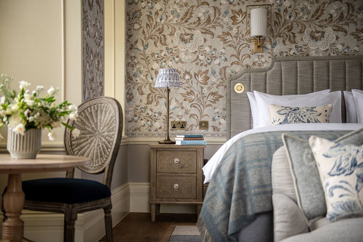 A bedroom with a bed, a nightstand with books, a lamp, a chair, and a vase with flowers.