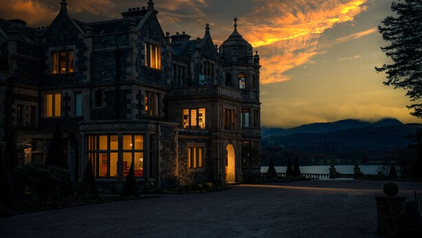 A large, historic building with multiple windows and a prominent entrance, surrounded by a landscaped area and a body of water in the background. - Langdale Chase Hotel (Windermere)