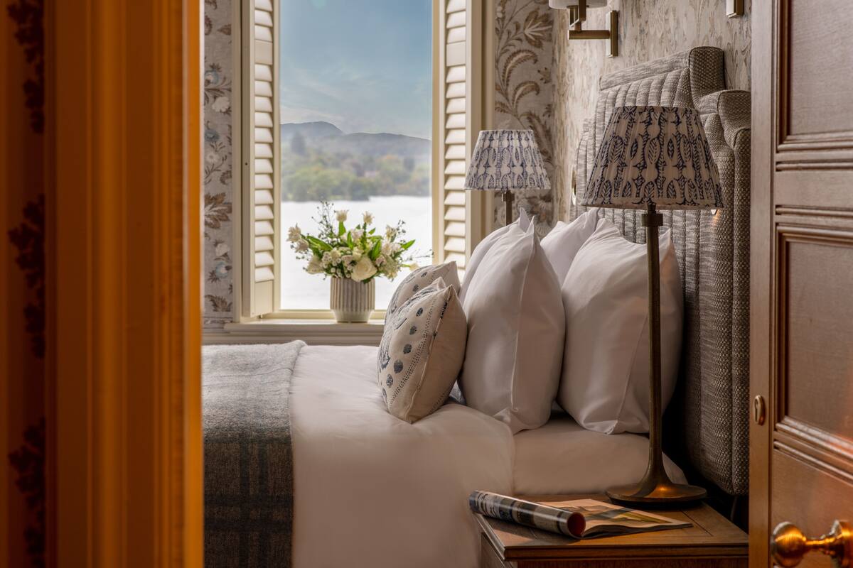A bedroom with a bed, two lamps, a window with a view of a lake, and a vase with flowers.
