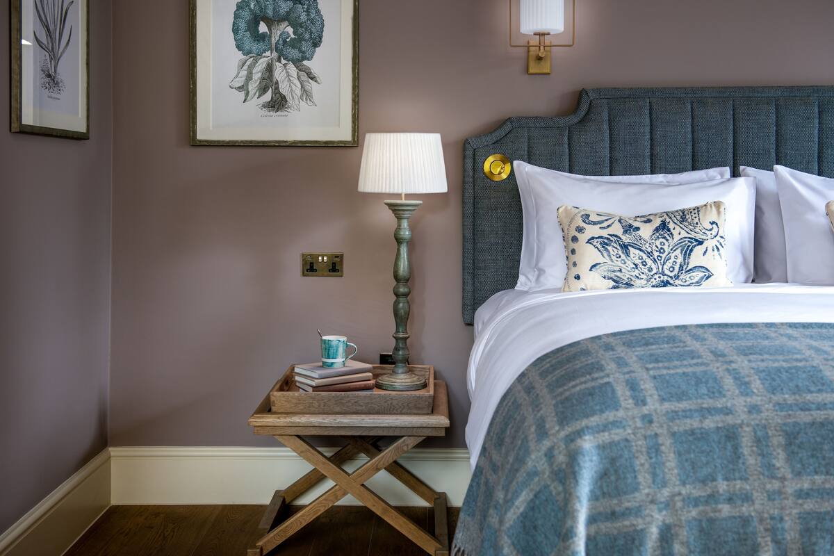 A neatly made bed with a blue patterned bedspread, a bedside table with a lamp, books, and a cup, and a framed botanical illustration on the wall.