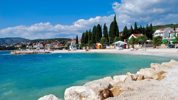 A coastal beach with rocky shoreline, clear turquoise water, and buildings in the background. - Villa Mediteran (Okrug)