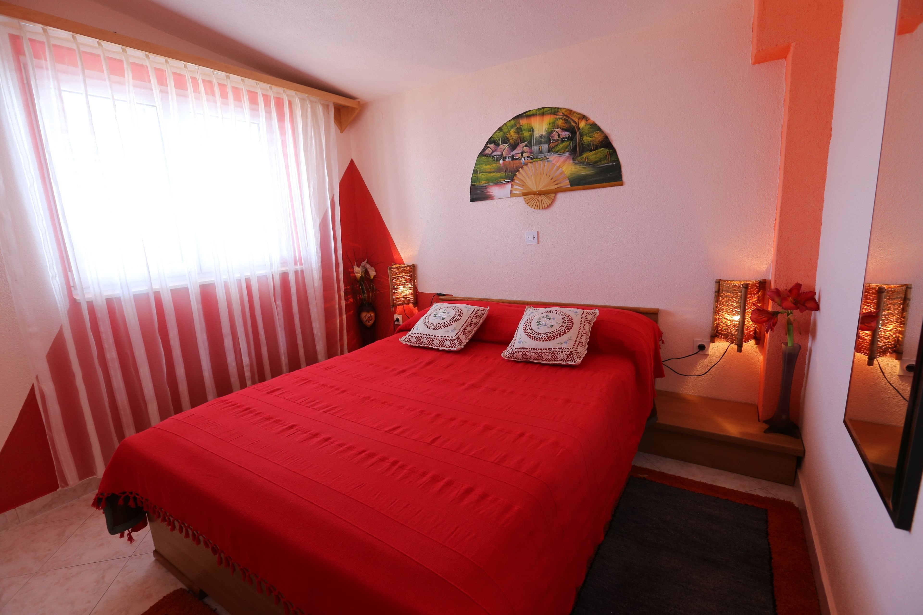 Une chambre avec une couvre-lit rouge, un tableau au mur et une fenêtre avec des rideaux.
