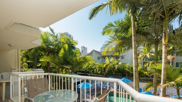 Uma varanda com mesa e cadeiras de vidro, cercada por palmeiras e vista para a área da piscina.