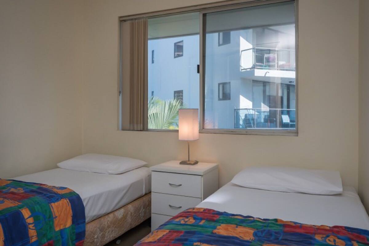 Habitación de hotel con dos camas, una mesita de noche con lámpara, una ventana con vistas a edificios y una puerta.