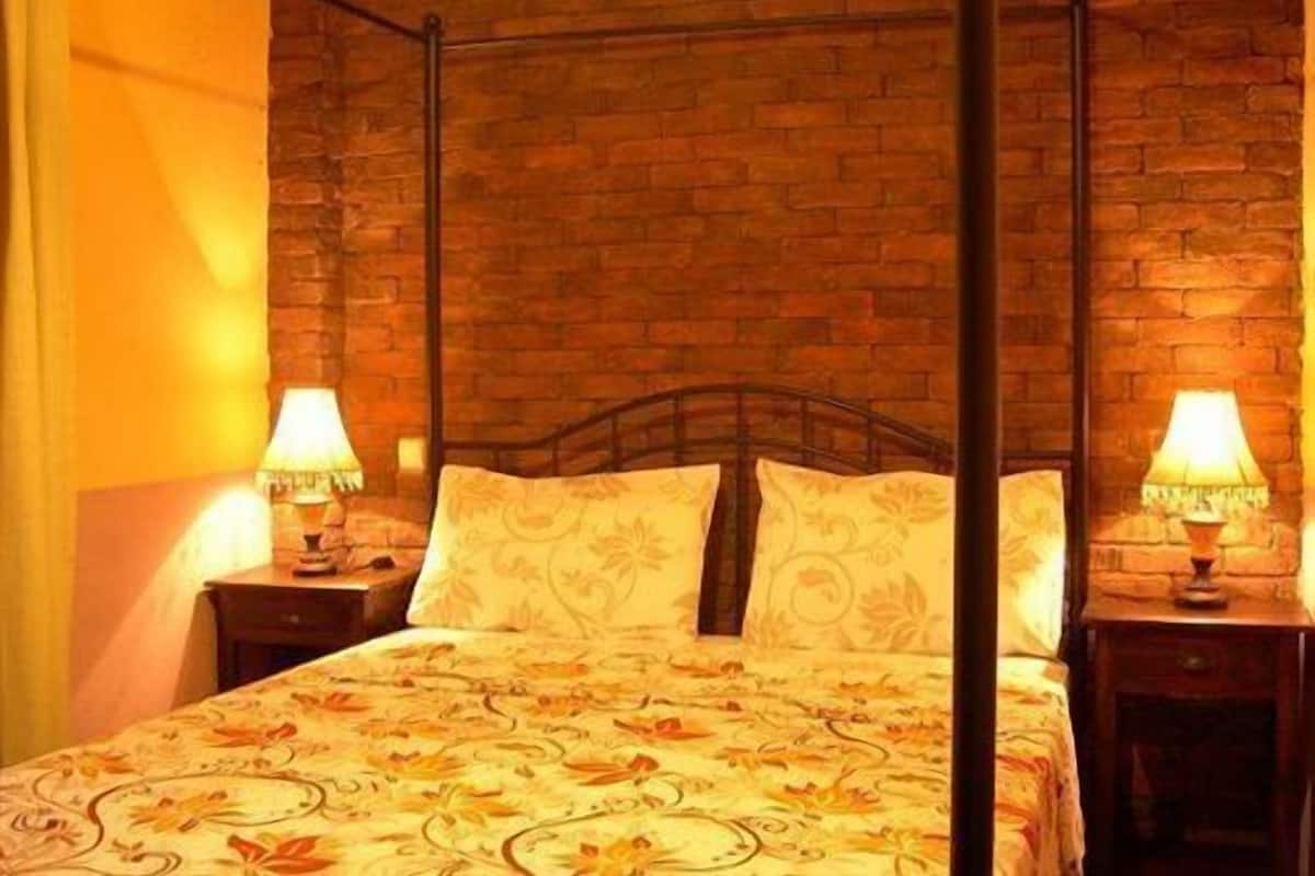 A room with a four-poster bed, two bedside tables with lamps, a brick wall, and a floral-patterned bedspread.