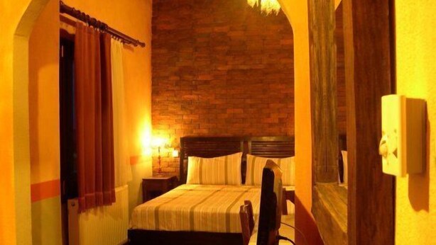 A cozy room with a brick wall, a bed with a wooden headboard, a bedside table, and a lamp. - Mythos Guesthouse (Kalabaka)