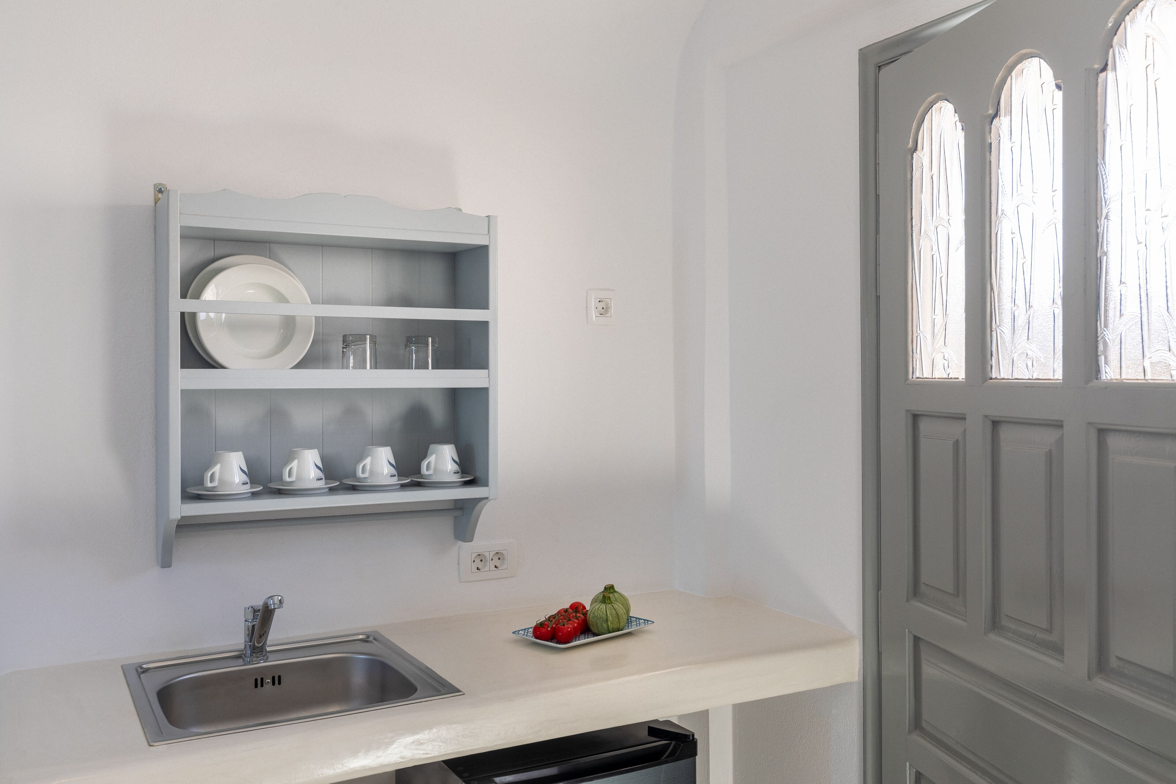 A kitchen with a built-in shelf holding plates and cups, a sink, and a small window with frosted glass.