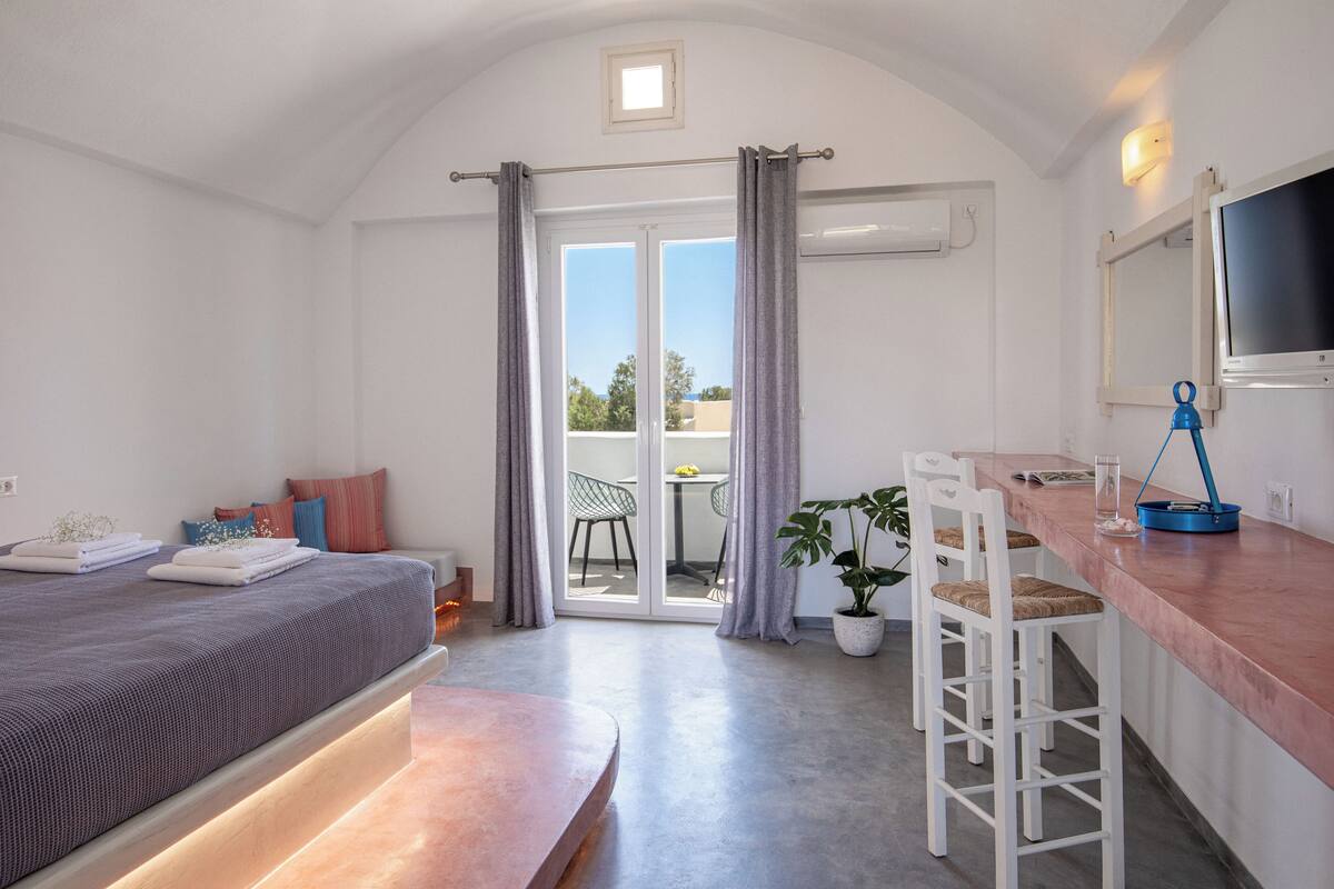 Quarto com cama, escrivaninha, cadeiras, planta em vaso e porta de vidro corrediça para uma varanda.