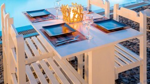 A poolside dining area with white chairs and tables, a clear pool, and a building with balconies in the background. -  Sea View Bay Hotel Santorini (Santorini)
