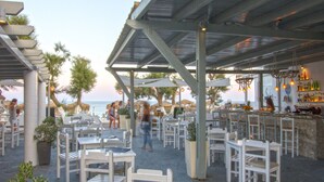 An outdoor dining area with white tables and chairs, a bar area with bottles, and a view of the ocean. -  Sea View Bay Hotel Santorini (Santorini)