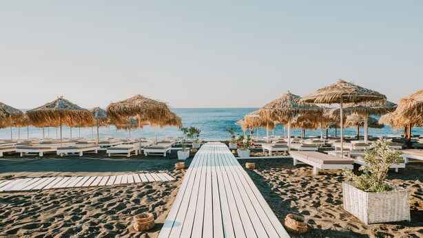 A beach with a wooden walkway leading to lounge chairs and thatched umbrellas. -  Sea View Bay Hotel Santorini (Santorini)