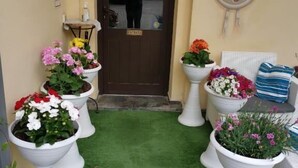 Entrada de hotel con césped artificial verde, macetas blancas con flores, una puerta marrón con un cartel y un letrero decorativo en la pared.