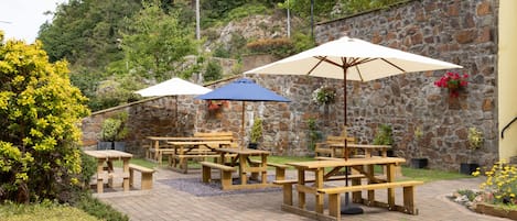 An outdoor patio with wooden picnic tables and umbrellas, surrounded by a stone wall and greenery.