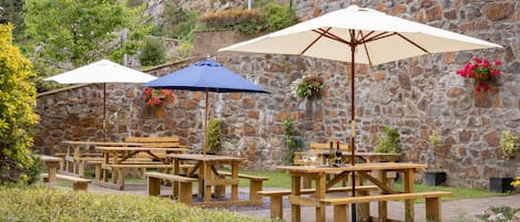 Outdoor wooden picnic tables with umbrellas, set against a stone wall with greenery and flowers.