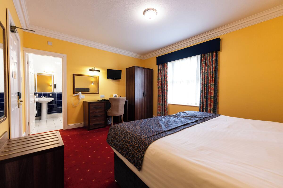 Une chambre d’hôtel comprenant un lit, un bureau, une chaise, une armoire, un miroir et une salle de bain visible à travers une porte ouverte.
