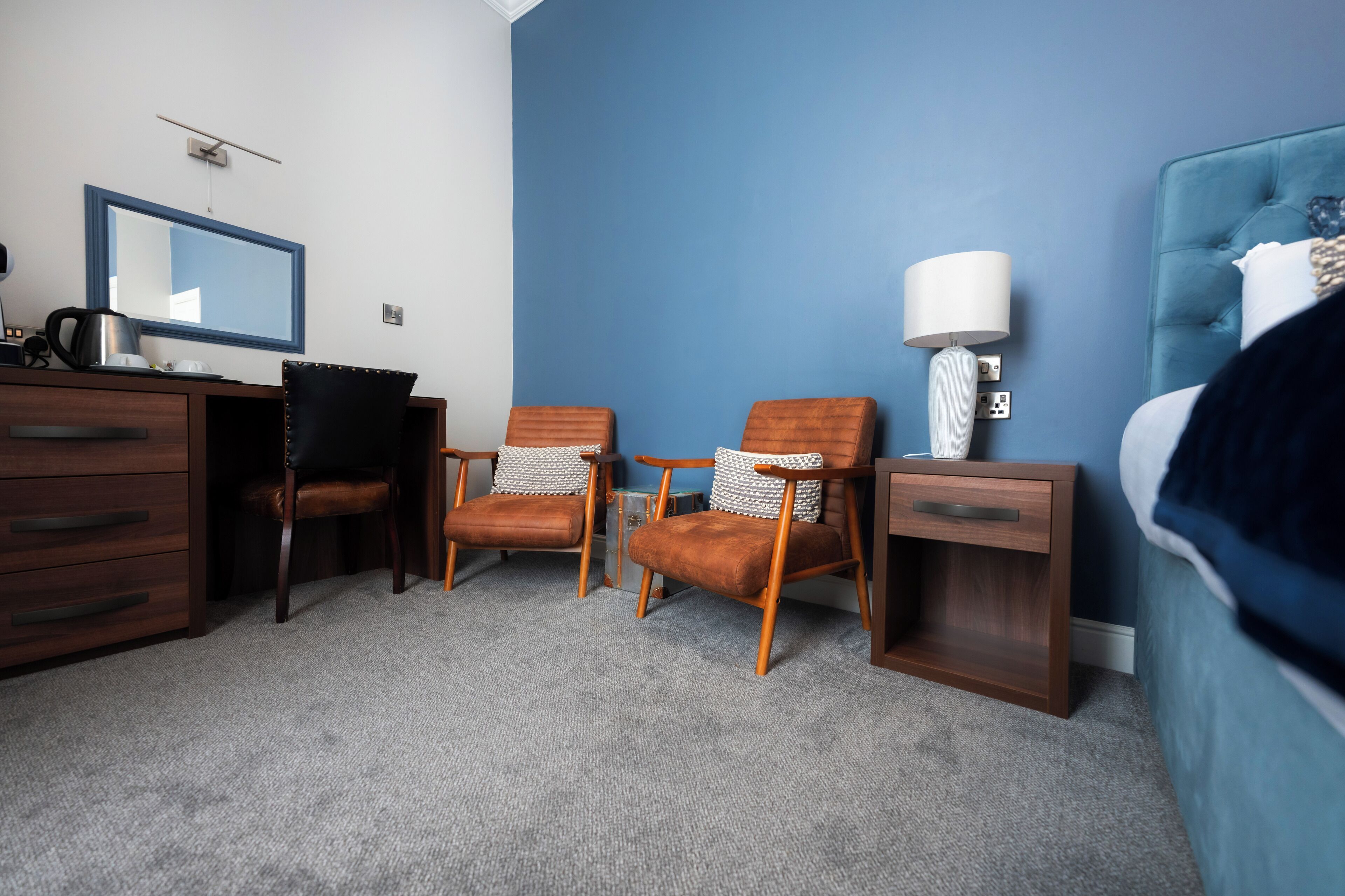 A hotel room with a blue accent wall, a bed, a desk with a chair, a nightstand with a lamp, and a mirror.