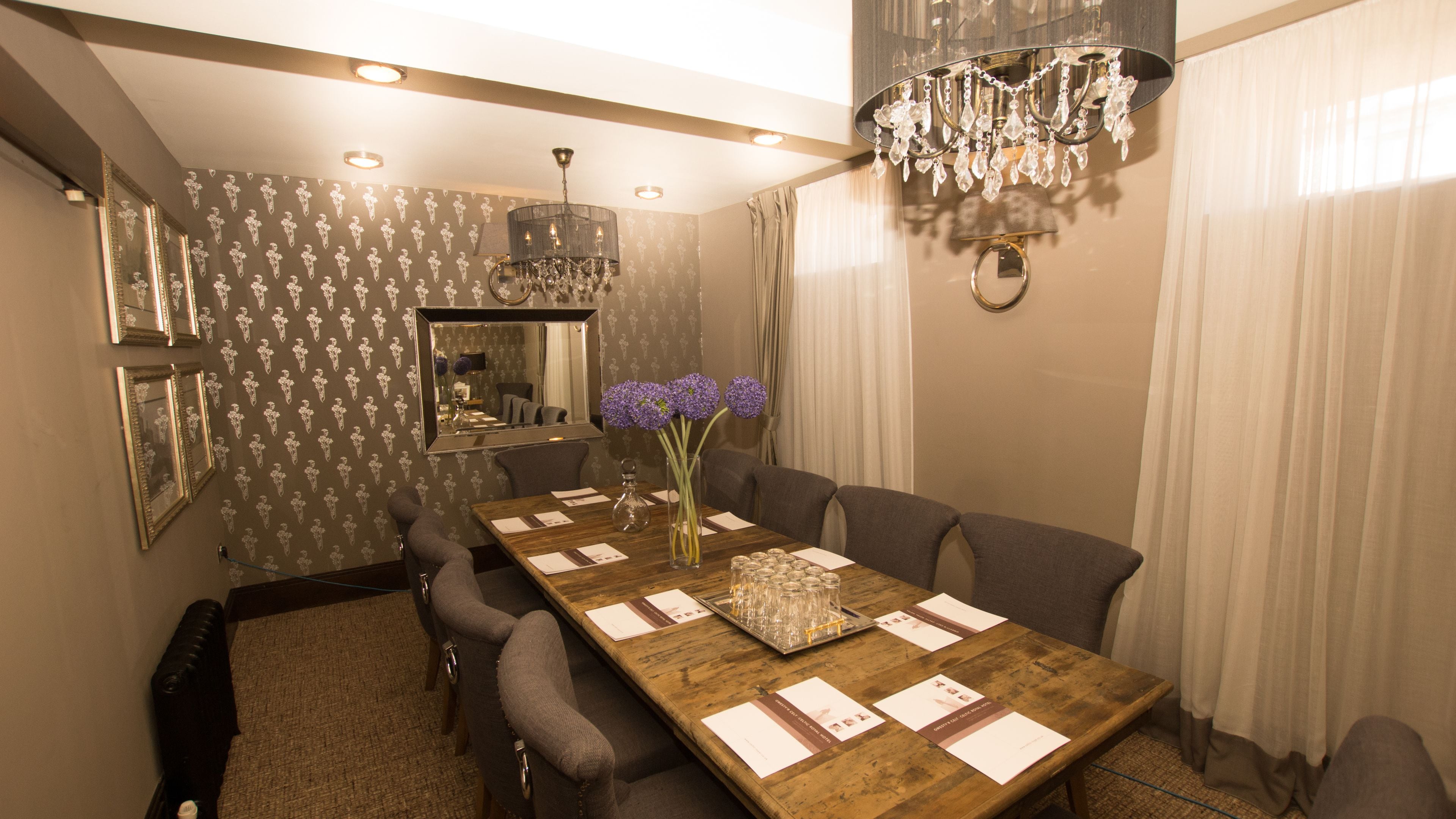 Comedor con una mesa de madera dispuesta para seis comensales, un jarrón con flores moradas y una araña.